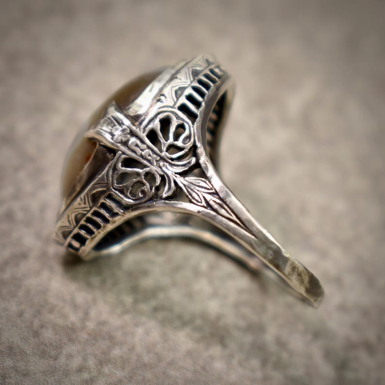 Puede incluir: Un anillo de plata con una piedra ovalada, lisa y de color marr&oacute;n claro engastada en una montura de metal decorativa. La banda y el engaste muestran intrincados patrones grabados y detalles de filigrana calada en los laterales.