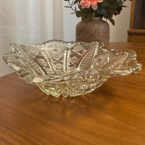May include: A clear, decorative glass bowl with a scalloped edge. The bowl features intricate starburst and geometric patterns. The bowl is sitting on a wooden table. A vase with flowers is in the background.