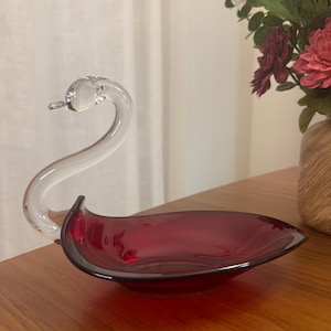 May include: A decorative glass swan dish with a ruby red bowl and a clear glass neck and head. The swan dish is sitting on a wooden surface. The swan dish is a decorative item.