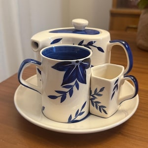 May include: A white ceramic tea set with blue floral designs. The set includes a teapot, two mugs, and a small square cup, all resting on a white saucer. The teapot and mugs have blue handles and rims.