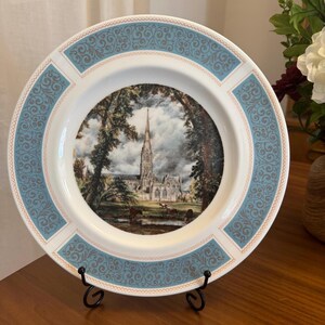 May include: A white ceramic plate with a blue and gold border. The center of the plate features a painting of a cathedral with trees in the foreground. The plate is on a black stand.