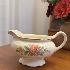 May include: Cream-colored ceramic gravy boat with a floral design featuring pink, yellow, and blue flowers. The boat has a handle and a scalloped base, with gold trim along the rim.