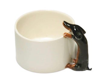 Puede incluir: Una taza de cerámica blanca con una figurita de dachshund unida al lado. El dachshund es negro y marrón con un pecho blanco y se agarra a la taza con sus patas.