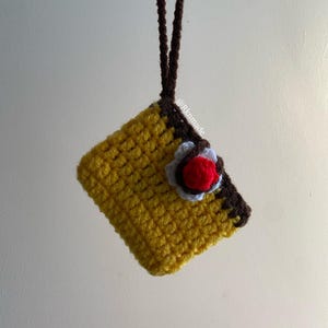 Könnte beinhalten: Handgefertigter gehäkelter Anhänger in quadratischer Form, gelb mit braunen Rändern und einer dekorativen Blume. Er hängt an einer braunen Kordel. Der Text "@Rkmade" ist auf der Seite des Anhängers zu sehen.