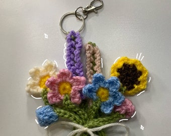 Könnte beinhalten: Ein gehäkelter Blumenstrauß-Schlüsselanhänger mit einem silbernen Schlüsselring. Der Strauß besteht aus verschiedenen Blumen in Rosa, Blau, Gelb und Lila mit grün gehäkelten Stielen und Blättern. Die Blumen sind mit einem weißen gehäkelten Band zusammengebunden.