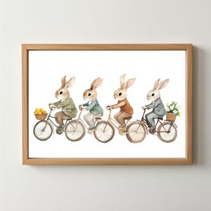May include: Watercolor print of four rabbits riding bicycles, each wearing a different outfit. The first rabbit carries a basket of yellow fruit. The print is framed in a light brown wooden frame.