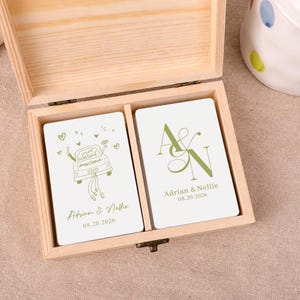 May include: A light-coloured wooden box containing two decks of white playing cards. One deck features a green line drawing of a car with the text "just married" and "Adrian & Nellie 08.20.2026". The other deck shows a green "A & N" monogram.