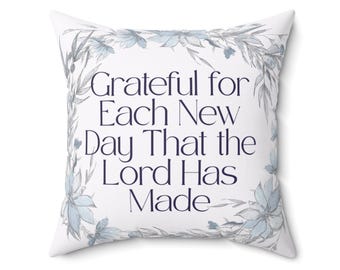 以下が含まれることがあります： 白い正方形のクッション。紺色の文字で「Grateful for Each New Day That the Lord Has Made」と書かれています。青とグレーの色合いの花輪で飾られています。