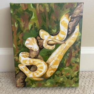 May include: A canvas painting depicts a yellow and white snake coiled around a brown tree branch. The background is a mix of green and brown, suggesting a jungle or forest setting. The snake's scales are detailed, and the artwork has a realistic style.