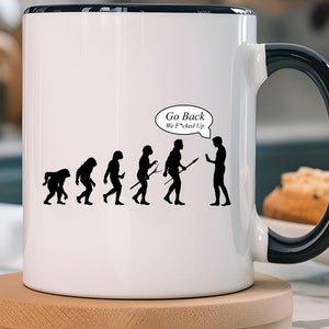 Puede incluir: Taza de cerámica blanca con asa y borde negros, con un gráfico de evolución en silueta negra y el texto "Go Back We F*cked Up" en un bocadillo. La taza está sobre un posavasos de madera.