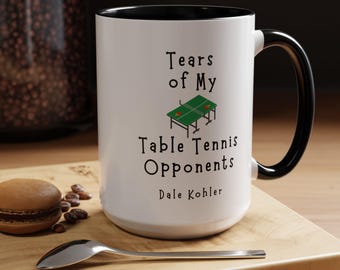 Könnte beinhalten: Weißer Keramikbecher mit schwarzem Innenraum und Henkel. Der Becher zeigt den Text "Tears of My Table Tennis Opponents" mit einer Grafik eines grünen Tischtennistisches. Der Name "Dale Kohler" ist darunter gedruckt.