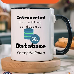 Może przedstawiać: Biały kubek ceramiczny z czarnym rantem i uchwytem. Kubek ma wzór z napisem "Introvert, ale chętny do dyskusji o bazie danych SQL" i imieniem "Cindy Hollman".