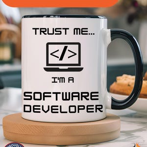 May include: White ceramic mug with a black handle and rim, featuring the text "TRUST ME... I'M A SOFTWARE DEVELOPER" and a laptop icon with code symbols. The mug sits on a wooden coaster. The words "Available In Other Colors" are at the top.