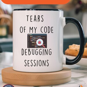Può includere: Tazza in ceramica bianca con manico e bordo neri, con la scritta "TEARS OF MY CODE DEBUGGING SESSIONS". La tazza include una grafica di uno schermo di computer con un bug e una lente d'ingrandimento. Il testo "Available In Other Colors" è in alto.