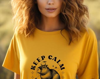 May include: Mustard yellow t-shirt with a graphic of a capybara wearing a fruit hat, surrounded by stars, and the text "KEEP CALM and CAPY ON". The shirt is a short-sleeved crew neck.