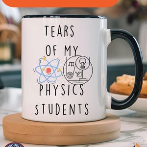 May include: White ceramic coffee mug with a black rim and handle. The mug has a black and white graphic of an atom, a light bulb, and a protractor with the text "Tears of my Physics Students".