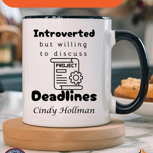 Puede incluir: Taza de cerámica blanca con borde y asa negros. La taza tiene texto negro que dice "Introvertido pero dispuesto a discutir" y "Plazos". Hay un gráfico de un documento con la palabra "Proyecto" y un engranaje. El texto "Cindy Hollman" está debajo del gráfico. La taza está sobre un posavasos de madera.