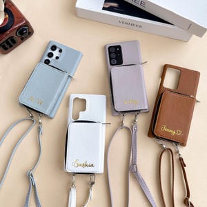 May include: Several leather phone cases in shades of blue, lilac, white, and brown. Each case has a zip-up wallet and a strap. Some cases are personalized with gold lettering, such as names or initials.