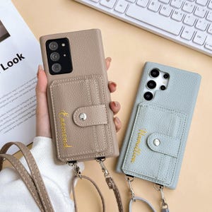 Peut inclure: Deux coques de téléphone avec porte-cartes et lanières. L'une est beige avec "Faemond" brodé en or, l'autre bleu clair avec "Hamilton" brodé en or. Les deux ont une fermeture à bouton-pression.