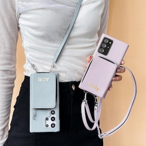 May include: Two phone cases with attached wallets. One is light blue with "AOR" in gold lettering, the other is lavender with "kelly" in gold lettering. Both have zippers and crossbody straps. The phone cases are designed to hold a smartphone.