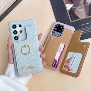 May include: Two phone cases are shown. One is light blue with a gold ring holder and the name "Maddy S" in gold script. The other is a light purple case with a brown wallet that holds a "NatWest Savings" card and a small mirror.