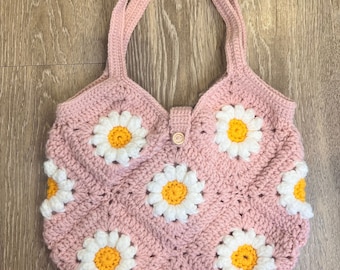 May include: A pink crocheted tote bag with white and yellow daisy flowers. The bag has two straps and a button closure.