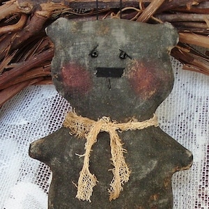 May include: A handmade, primitive-style bear doll with a distressed, dark gray fabric. The bear has rosy cheeks, a black stitched mouth, and a burlap scarf. The doll is a rustic, country-themed decorative item.