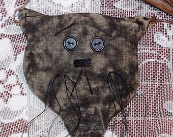 Op de afbeelding: Een grijze stoffen kattenpop met zwarte wollen snorharen en bruine knoopjesogen. De pop is bevestigd aan een stok met bruin draad en belletjes.