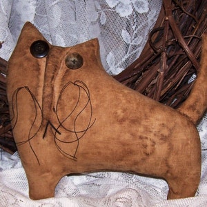 May include: A brown fabric cat with black embroidered whiskers and two brown buttons for eyes. The cat is sitting on a white lace fabric background.