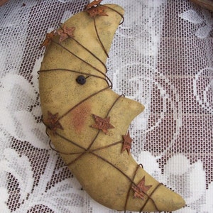 May include: A rustic, handmade crescent moon ornament with a wire frame and rusty star accents. The moon is made of fabric and has a distressed, aged look.