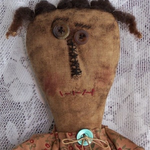 Könnte beinhalten: Eine handgemachte Stoffpuppe mit braunen Wollhaaren, einem braunen Stoffgesicht und zwei unterschiedlichfarbigen Knöpfen als Augen. Die Puppe trägt ein braunes Kleid mit Blumenmuster und einen hellblauen Knopf in der Taille.