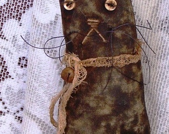 Op de afbeelding: Een handgemaakte primitieve stijl kattenpop met een bruin en zwart stoffen lichaam, een jute strik en een kleine bel. De kat heeft zwarte draad snorharen en grote bruine ogen.