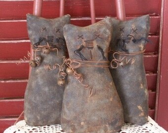 Op de afbeelding: Drie handgemaakte stoffen kattenpoppen met verweerd bruin stof, zwarte borduurdetails en kleine metalen belletjes die met draad zijn bevestigd.