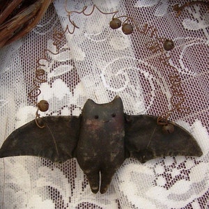 May include: A handmade fabric bat with brown wings and a black body, hanging from a wire with bells. The bat is hanging against a white lace background.