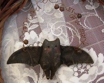 May include: A handmade fabric bat with brown wings and a black body, hanging from a wire with bells. The bat is hanging against a white lace background.