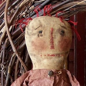 Könnte beinhalten: Eine handgemachte Stoffpuppe mit roten Wollhaaren, einem gestickten Gesicht und einem rot-weiß karierten Kleid. Die Puppe trägt eine Sicherheitsnadel und eine Glocke.
