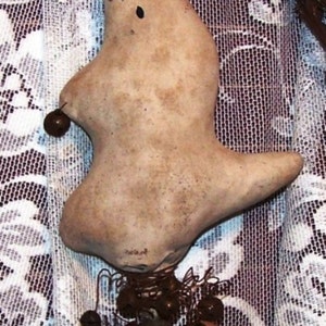 Pode incluir: Uma figurinha de fantasma feita à mão com um acabamento branco desgastado e olhos pretos. O fantasma está preso a um pau com sinos marrons pendurados.