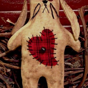 May include: A handmade rag doll bunny with long ears, brown fabric, and a red plaid heart sewn on the front. The doll has two black buttons for eyes and is sitting on a woven basket.