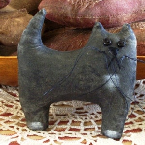 May include: A handmade, gray cat-shaped stuffed animal with button eyes and stitched whiskers. The cat has a curved tail and white accents on its feet. The plush toy is displayed on a lace doily.