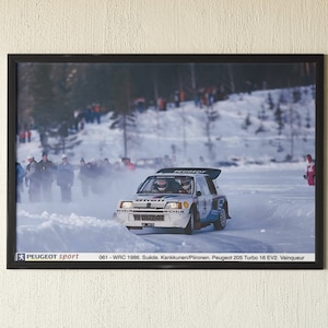 Puede incluir: Una fotografía enmarcada de un coche de rally blanco conduciendo por una pista cubierta de nieve. El coche es un Peugeot 205 Turbo 16 EV2, y el texto debajo de la imagen dice "061-WRC 1986. Suide. Kankkunen/Piironen. Peugeot 205 Turbo 16 EV2. Vainqueur."