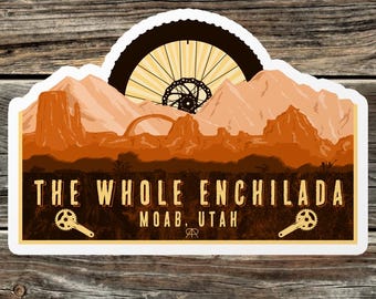 May include: A sticker with a mountain landscape and a bicycle wheel in the background. The text "THE WHOLE ENCHILADA" is at the top, with "MOAB, UTAH" below it. There are two bicycle chainrings on either side of the text.