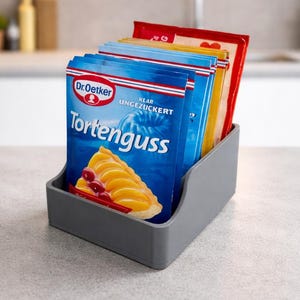 Puede incluir: Un organizador de plástico gris que contiene varios paquetes de Dr. Oetker Tortenguss. Los paquetes son azules, rojos y amarillos, con el paquete azul que muestra una tarta de frutas. El organizador está sobre una encimera gris.