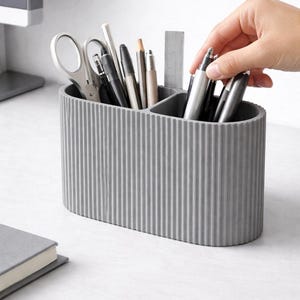 Könnte beinhalten: Ein grauer, geriffelter Schreibtisch-Organizer gefüllt mit Stiften, Bleistiften, Scheren und einem Lineal. Der oval geformte Organizer ist in drei Abschnitte unterteilt. Eine Hand greift in den Organizer, um einen Stift zu greifen. Der Organizer steht auf einer weißen Oberfläche.