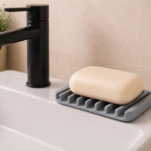 Könnte beinhalten: Ein hellbeige Seifenstück liegt auf einer grauen Seifenschale. Ein schwarzer Wasserhahn befindet sich über einem weißen Waschbecken. Eine kleine Topfpflanze und zwei braune Flaschen sind im Hintergrund.