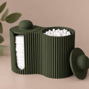 Può includere: Un organizer da bagno verde oliva scuro, a coste, con due scomparti e forma arrotondata. Un lato presenta un'apertura verticale riempita di dischetti di cotone bianchi, mentre l'altro è un contenitore aperto riempito di bastoncini cotonati.