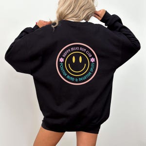 Puede incluir: Sudadera negra con un gráfico de cara sonriente rosa, amarillo y azul. El gráfico dice "Happy Miles Run Club, Positive Mind & Negative Splits".