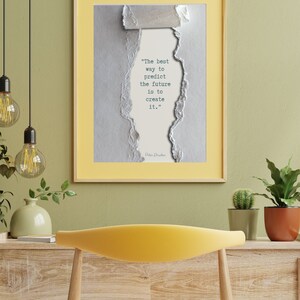 Könnte beinhalten: Ein gerahmter Druck mit gelbem Rahmen und einem Zitat von Peter Drucker. Das Zitat "Der beste Weg, die Zukunft vorherzusagen, ist, sie zu gestalten" wird auf einem zerrissenen Papierhintergrund angezeigt. Der Druck befindet sich über einem gelben Stuhl.