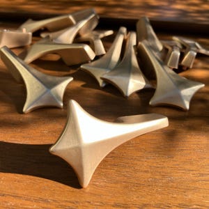 Pode incluir: Uma coleção de puxadores dourados em forma de estrela para armários. Cada puxador apresenta um design de estrela de quatro pontas com uma pega curva. Os puxadores estão dispostos sobre uma superfície de madeira, mostrando o seu acabamento metálico e forma geométrica.