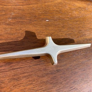 May include: A mid-century modern starburst cabinet pull with a gold finish. The hardware is made of metal and is set against a wooden surface. The starburst design has four elongated points.