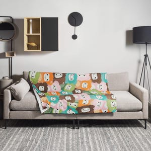 May include: A soft, plush throw blanket featuring a colorful pattern of cartoon faces. The blanket is displayed on a light gray couch with a throw pillow.